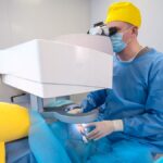 A surgeon wearing a blue surgical gown, a yellow cap, and white latex gloves looks into a machine to perform eye surgery.