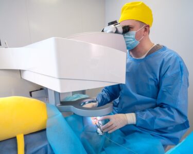 A surgeon wearing a blue surgical gown, a yellow cap, and white latex gloves looks into a machine to perform eye surgery.