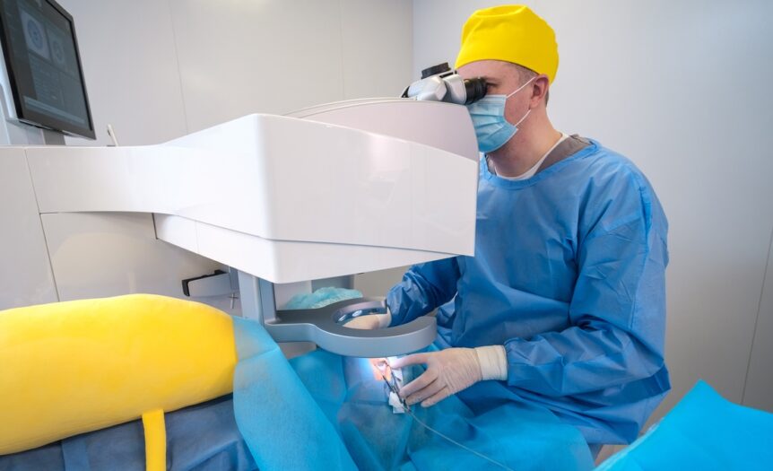A surgeon wearing a blue surgical gown, a yellow cap, and white latex gloves looks into a machine to perform eye surgery.