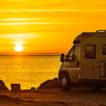 An RV parked up on a small cliff. It's overlooking the ocean with the sunset in the sky adding gold to everything.