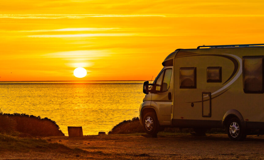 An RV parked up on a small cliff. It's overlooking the ocean with the sunset in the sky adding gold to everything.