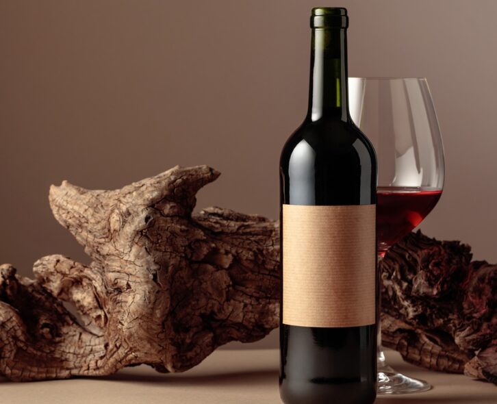 A wine bottle and glass of red wine in front of a piece of weathered wood on a brown background.