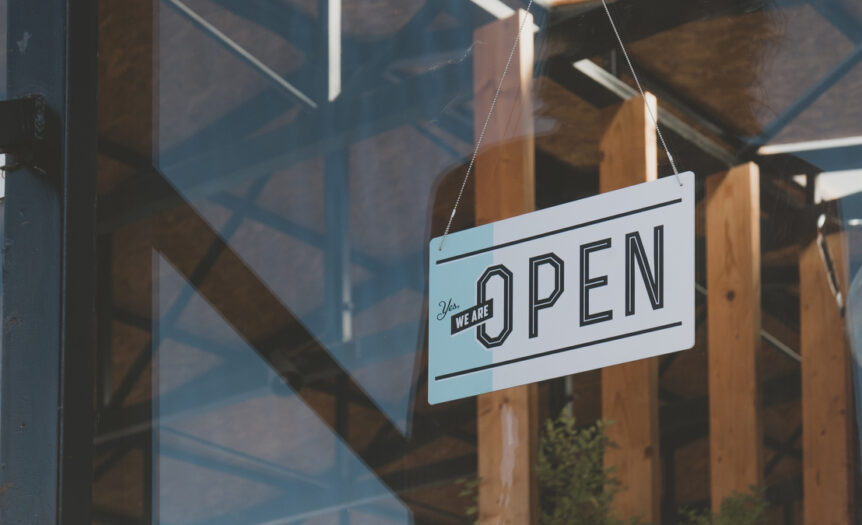 A window to a business with a white sign that says “Yes, we are open” on it in black lettering.