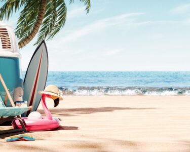 A sandy beach on a clear day. To the left is a blue vintage camper van, a beach chair, a guitar, and some flotation devices.