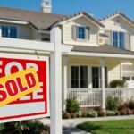 A yellow sticker with red lettering reading “SOLD” covers a “FOR SALE” sign in front of a house.