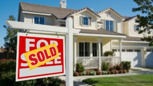 A yellow sticker with red lettering reading “SOLD” covers a “FOR SALE” sign in front of a house.