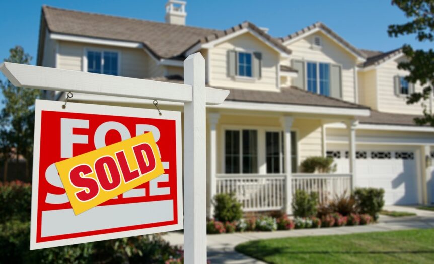 A yellow sticker with red lettering reading “SOLD” covers a “FOR SALE” sign in front of a house.