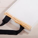 A white fire blanket with black tabs sits on a light-colored wooden surface. The blanket is folded.