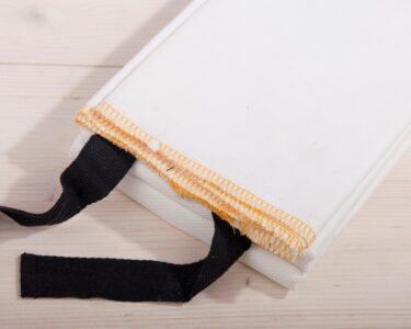 A white fire blanket with black tabs sits on a light-colored wooden surface. The blanket is folded.