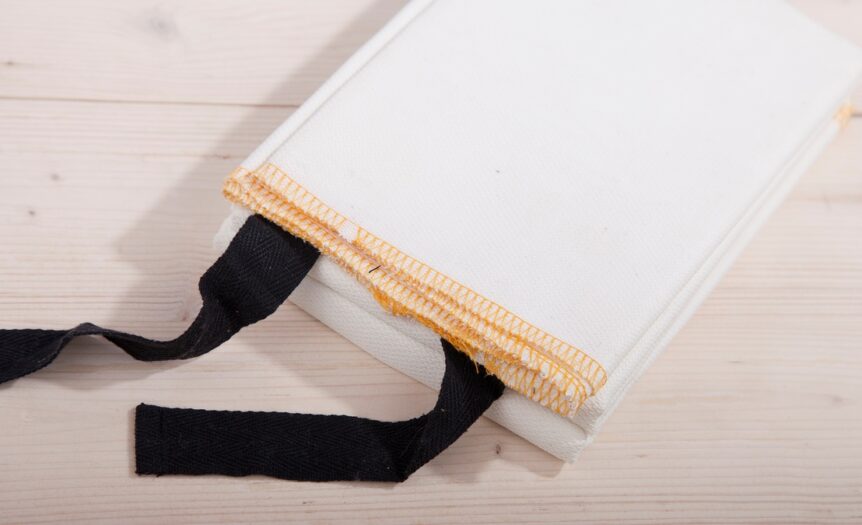 A white fire blanket with black tabs sits on a light-colored wooden surface. The blanket is folded.