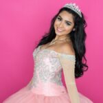 A smiling young woman with wavy black hair wearing a light pink dress with silver embroidery and beading on the bodice.