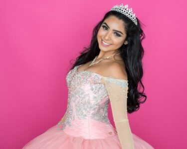 A smiling young woman with wavy black hair wearing a light pink dress with silver embroidery and beading on the bodice.