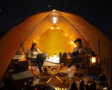 A young man and woman sitting on chairs and typing on laptops inside of a large tent covered in fairy lights.