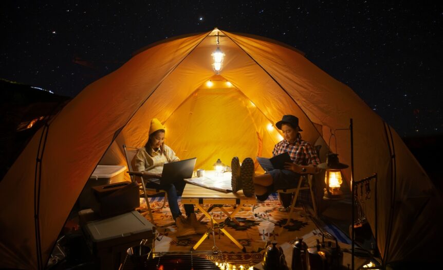 A young man and woman sitting on chairs and typing on laptops inside of a large tent covered in fairy lights.