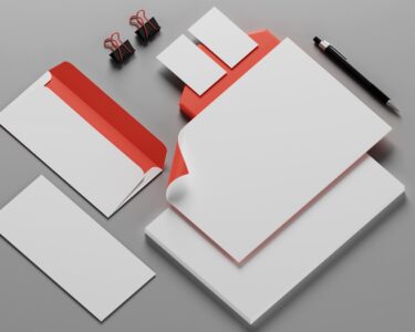A gray desk covered in white and orange-colored business stationery, including paper, envelopes, binder clips, and a pen.