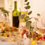 A close-up of a dining room table with wine glasses, a bowl of salad, cherry tomatoes, and a vase with eucalyptus.