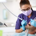 A female in purple scrubs, gloves, and a face mask using tools to clean a male patient's mouth in a dentist's office.