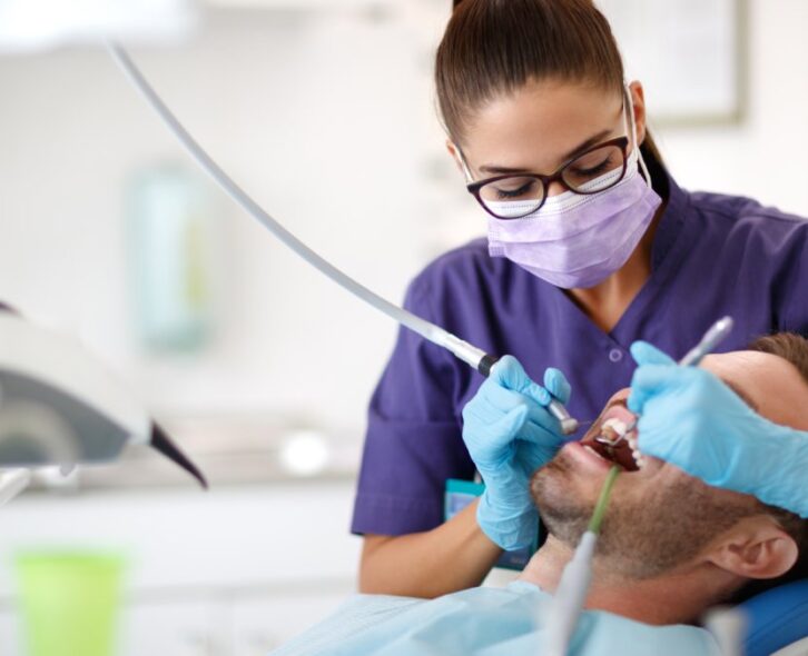 A female in purple scrubs, gloves, and a face mask using tools to clean a male patient's mouth in a dentist's office.