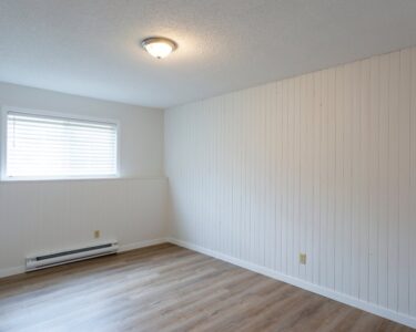 An empty room with white paneled walls, wood flooring, a window, an overhead light, and a white baseboard heater.