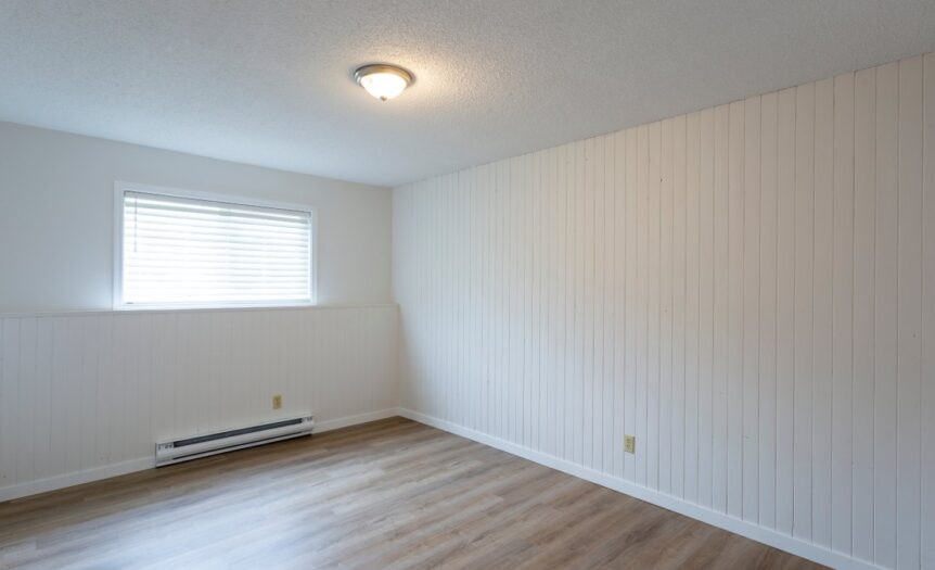 An empty room with white paneled walls, wood flooring, a window, an overhead light, and a white baseboard heater.