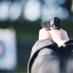 A close-up view shows a person wearing a black jacket holding a small, black gun and aiming at a target.