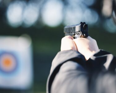 A close-up view shows a person wearing a black jacket holding a small, black gun and aiming at a target.