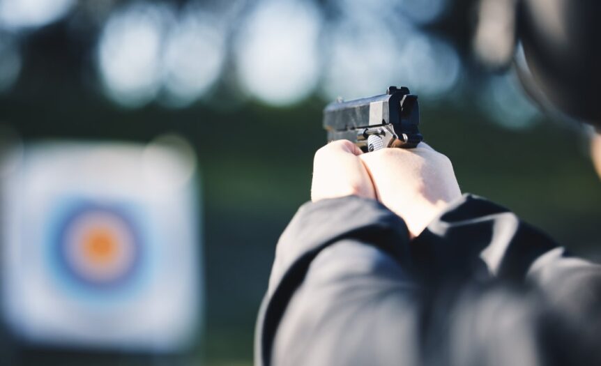 A close-up view shows a person wearing a black jacket holding a small, black gun and aiming at a target.