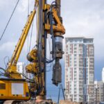 A hydraulic drilling rig is installing bored piles with a casing string on a construction site next to skyscrapers.