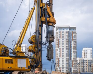 A hydraulic drilling rig is installing bored piles with a casing string on a construction site next to skyscrapers.