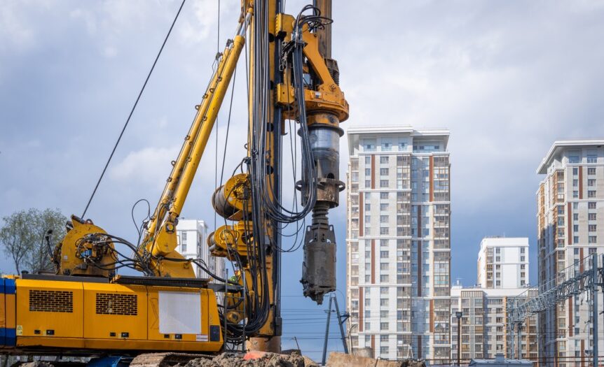 A hydraulic drilling rig is installing bored piles with a casing string on a construction site next to skyscrapers.