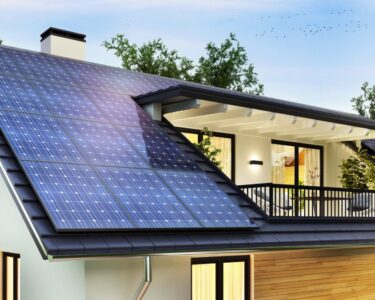 A home with black shingles has a second-story balcony and solar panels on the roof. The home is surrounded by green foliage.