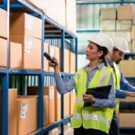 Two workers in safety vests and hard hats inside a warehouse. One scans boxes while the other works on a laptop.