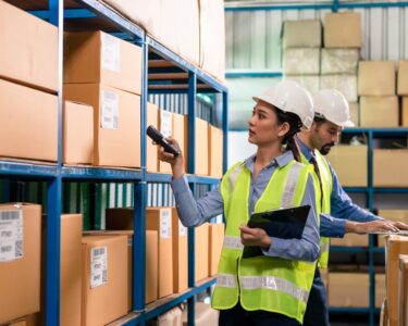 Two workers in safety vests and hard hats inside a warehouse. One scans boxes while the other works on a laptop.