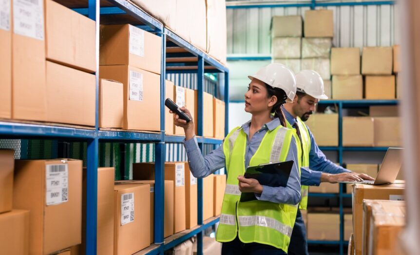 Two workers in safety vests and hard hats inside a warehouse. One scans boxes while the other works on a laptop.