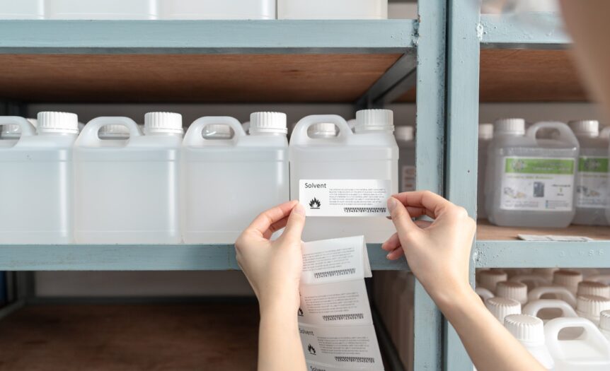A person placing a safety/warning label onto a clear bottle holding solvent based chemicals for storage.