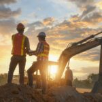 Two construction workers wearing orange safety vests stand on a pile of dirt next to a large excavator.