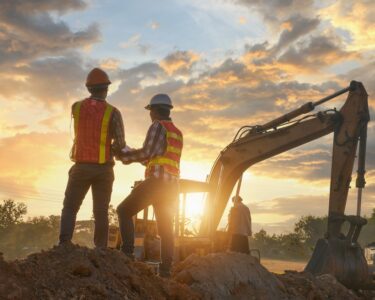Two construction workers wearing orange safety vests stand on a pile of dirt next to a large excavator.
