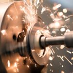 A close-up of a lathe grinder's tailstock spindle, spinning rapidly and producing heat sparks on a jaw chuck.