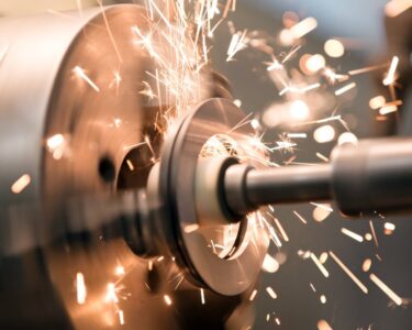A close-up of a lathe grinder's tailstock spindle, spinning rapidly and producing heat sparks on a jaw chuck.