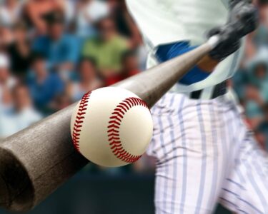 A close-up of playing using a dark brown bat to connect with a pristine white baseball with red stitching.