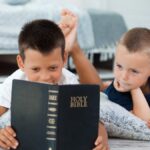 Two young boys laying next to each other on the floor reading the Bible. There is a pallet bed behind them.