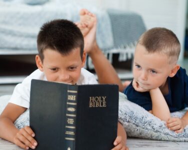 Two young boys laying next to each other on the floor reading the Bible. There is a pallet bed behind them.