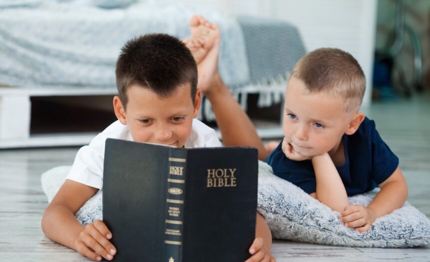 Two young boys laying next to each other on the floor reading the Bible. There is a pallet bed behind them.