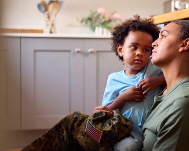 A woman sits on wood flooring against blue cabinetry, holding a kid with a camo cap and an American flag on his knees.