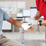 A person in a red shirt carefully applies a bandage to a man's hand who is sitting on a chair, offering first aid care.