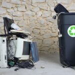 Old computer equipment and electronics are piled on the ground and fill a recycling bin next to a broom.