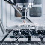 A close-up view shows a machine drilling into a piece of metal. Water is spraying all around the metal and the machine.