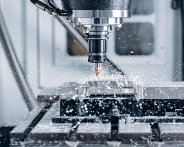 A close-up view shows a machine drilling into a piece of metal. Water is spraying all around the metal and the machine.