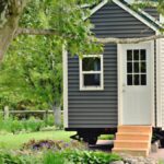 The exterior of a tiny home on wheels with gray siding, white trim, wooden front steps, and a white front door.