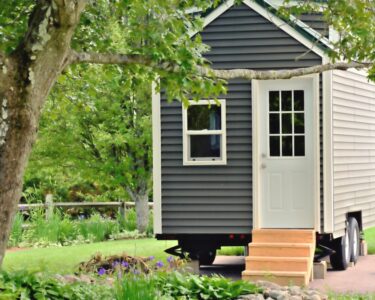 The exterior of a tiny home on wheels with gray siding, white trim, wooden front steps, and a white front door.
